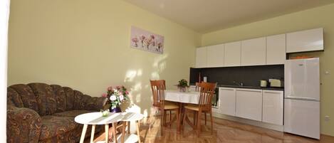 Una sala de estar con sofá, una mesa de comedor con sillas, una cocina con armarios blancos y un refrigerador.