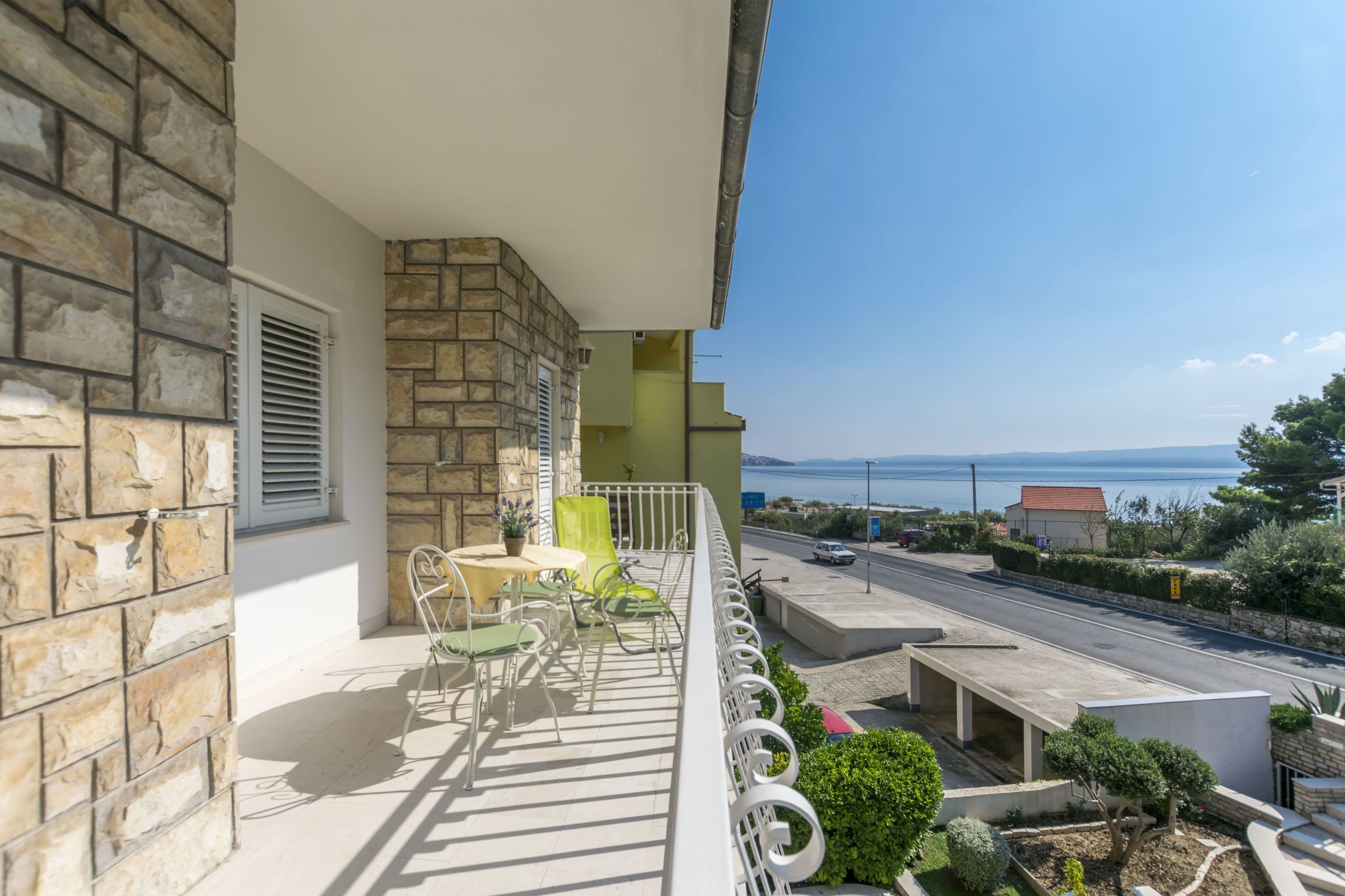 A balcony with stone walls, a table with chairs, and a view of a road and sea.