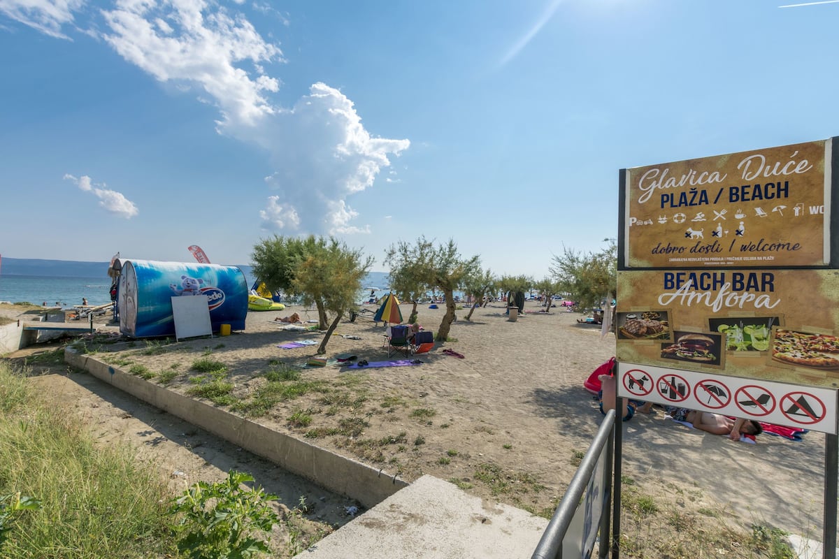 A beach area with a sign indicating 'Glavica Duće Plaza / Beach' and 'Beach Bar Amfora'.