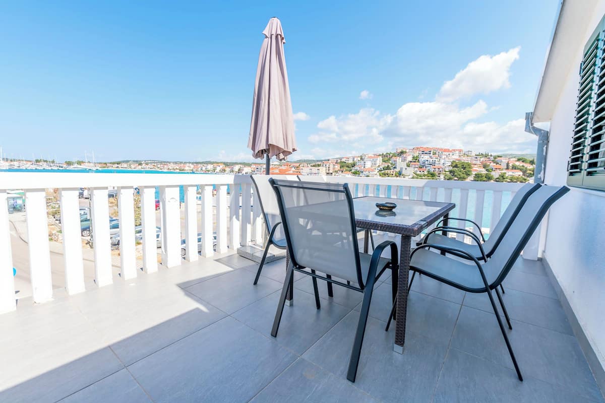 Un balcon avec une table et des chaises, une vue sur une ville côtière et un ciel bleu dégagé.