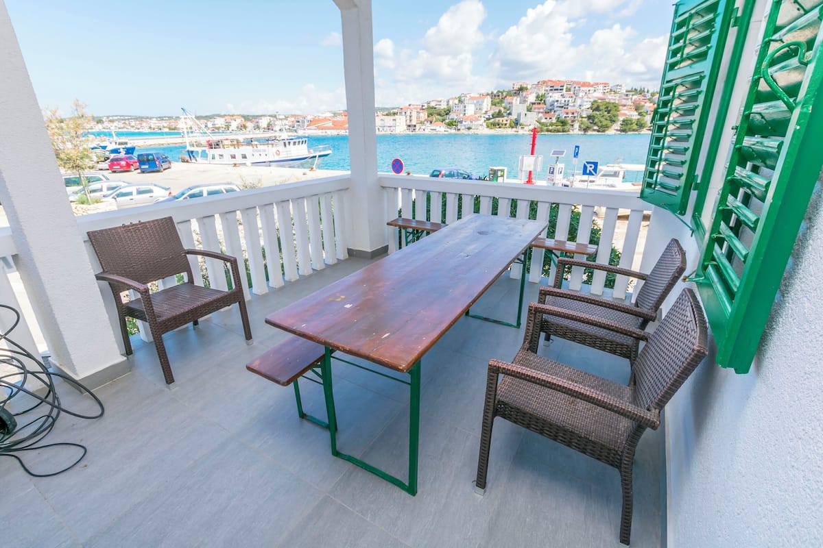 Un balcon avec une table en bois et des chaises en osier, donnant sur une marina où sont amarrés des bateaux et sur une ville côtière.