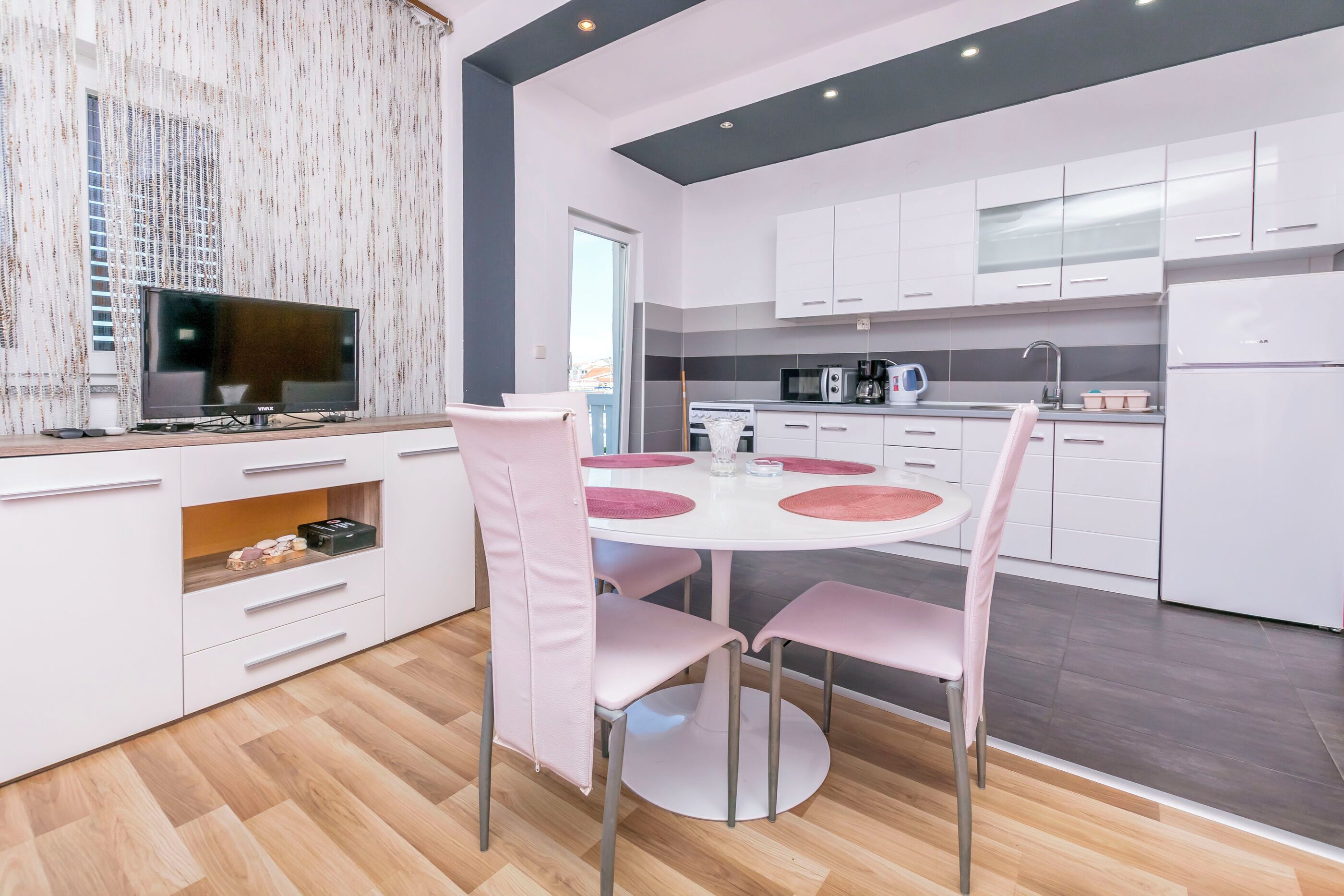 A modern kitchen with white cabinets, a dining table with pink chairs, and a flat-screen TV on a stand.