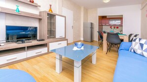 A modern living room with a glass coffee table, a blue sofa, and a TV on a wooden stand. - Apartments Olivera (Rogoznica)