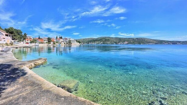 A coastal scene with clear water, a concrete walkway, and buildings in the background. - Apartments Olivera (Rogoznica)