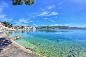 A coastal scene with clear water, a concrete walkway, and buildings in the background. - Apartments Olivera (Rogoznica)