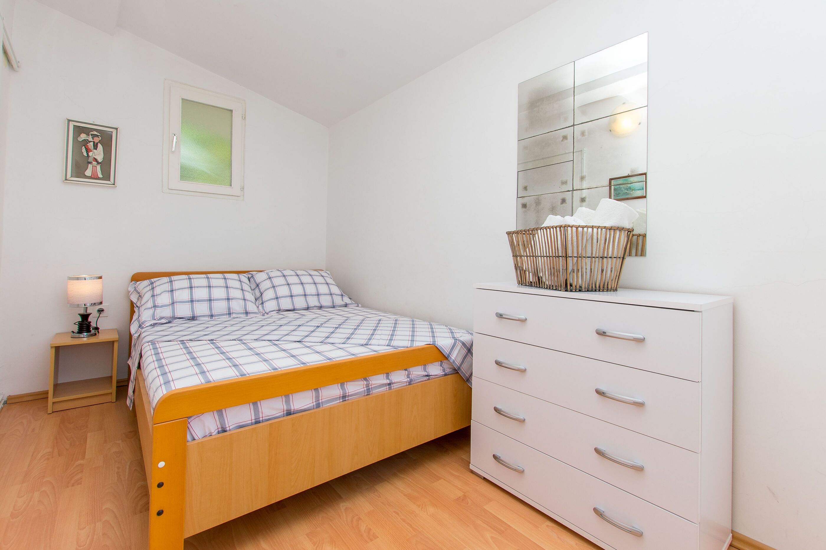 Une chambre avec un lit en bois, une commode blanche, une table de chevet avec une lampe et un grand miroir au mur.
