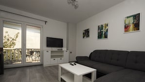 Un soggiorno moderno con un divano grigio, un tavolino da caffè bianco e una TV a schermo piatto su un mobile bianco. Due quadri decorano la parete, e una porta vetrata scorrevole dà accesso a una zona esterna con tavolo e sedie da patio.