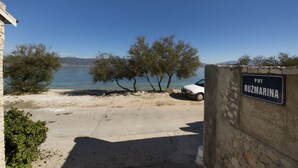Una strada costiera con un'auto bianca parcheggiata vicino a un muro di pietra, alberi e un cielo azzurro limpido. Un cartello recita "PUT RUŽMARINA".