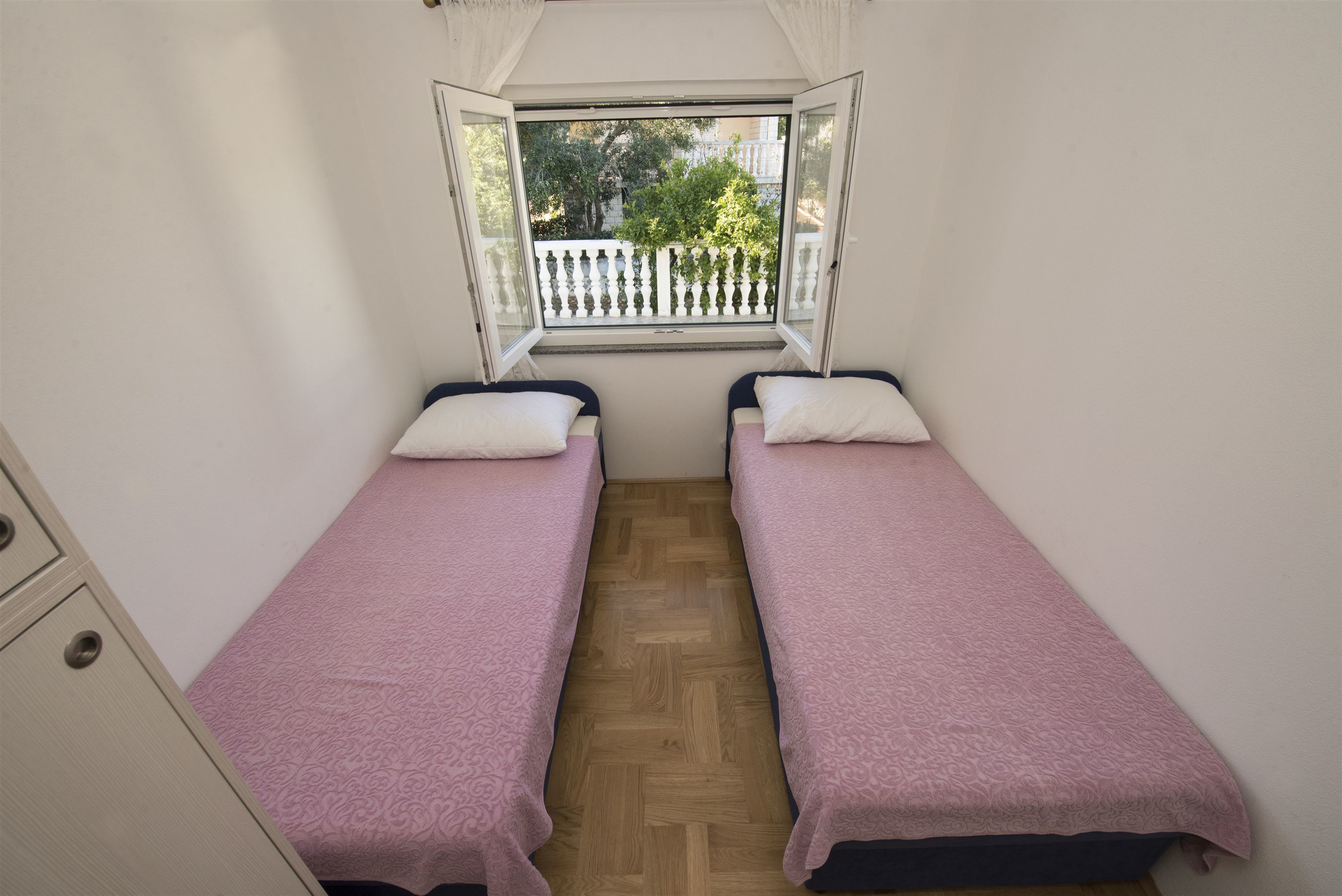A small, single-room accommodation with two single beds, a wooden floor, a window with a view of a balcony, and a white wardrobe.