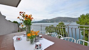 A table set with a vase of orange flowers, a metal pot, and a white tablecloth on a balcony overlooking a body of water and hills. - Apartment Jakov (Okrug Donji)