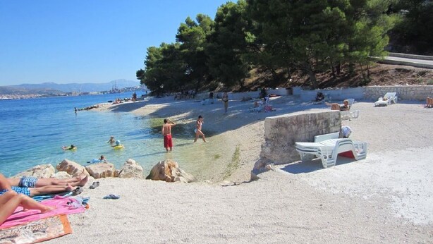 A beach with clear blue water, people swimming and sunbathing, and a rocky outcrop with a small white structure. - Apartment Jakov (Okrug Donji)