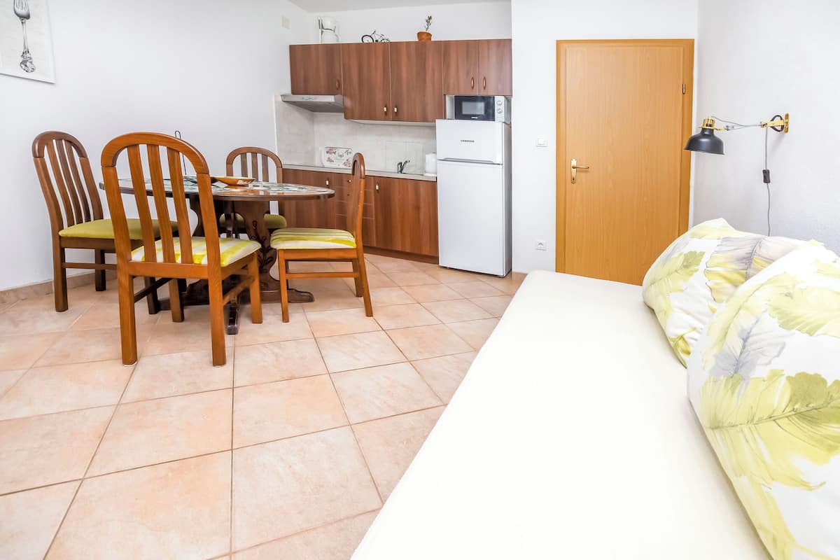 A compact kitchen with wooden cabinets, a refrigerator, and a microwave. There is a dining table with chairs and a bed with a floral bedspread.