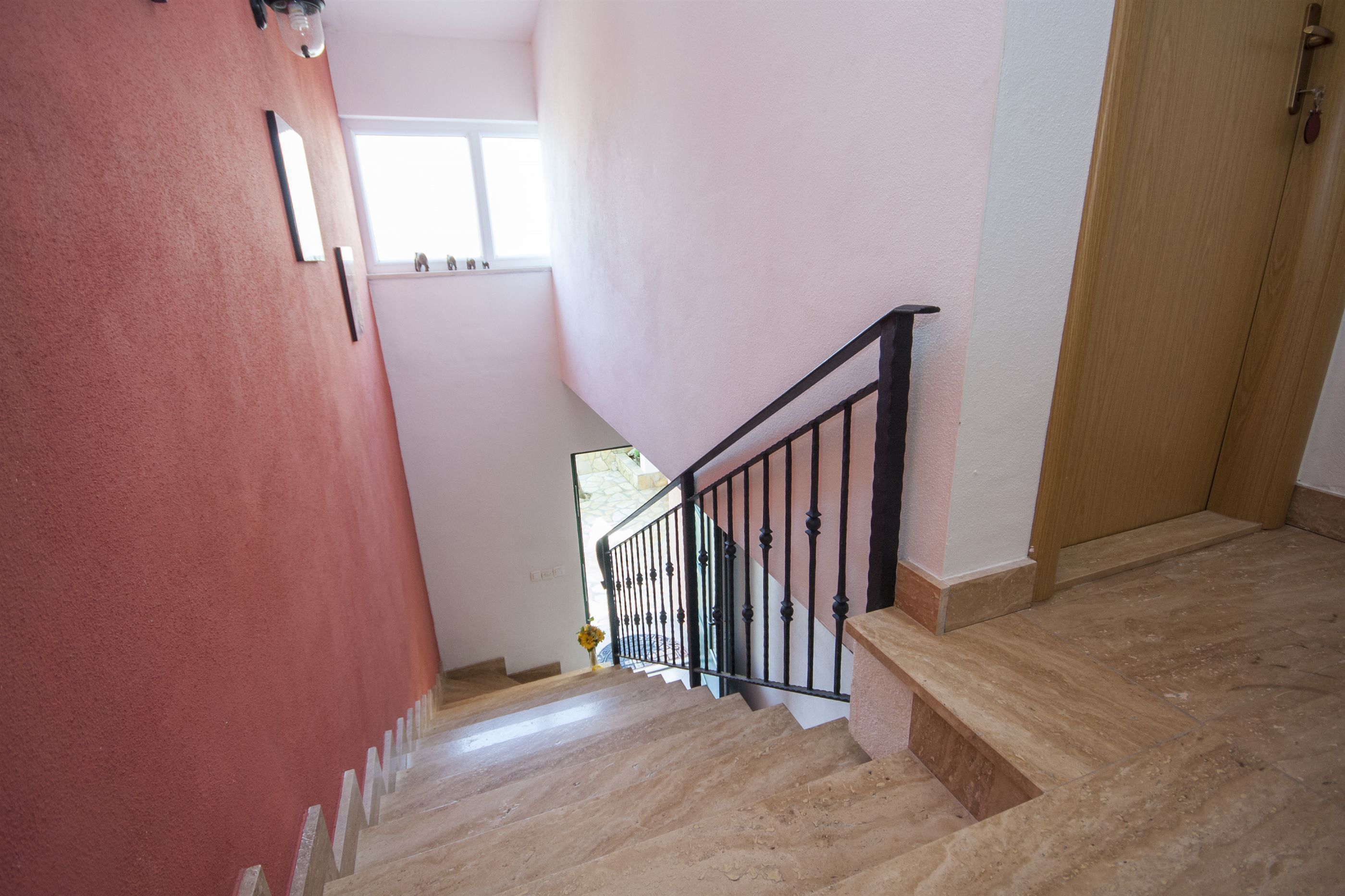 A staircase with a wooden handrail and a black metal railing, leading to a room with a pink wall and a window.