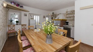 A dining area with a wooden table, chairs, and a vase of flowers. There is a kitchen in the background with shelves and appliances. - House Jasna (Igrane)