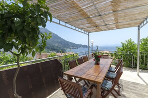Un espace de restauration en plein air avec une table et des chaises en bois, une pergola et une vue sur un paysage montagneux et la mer.