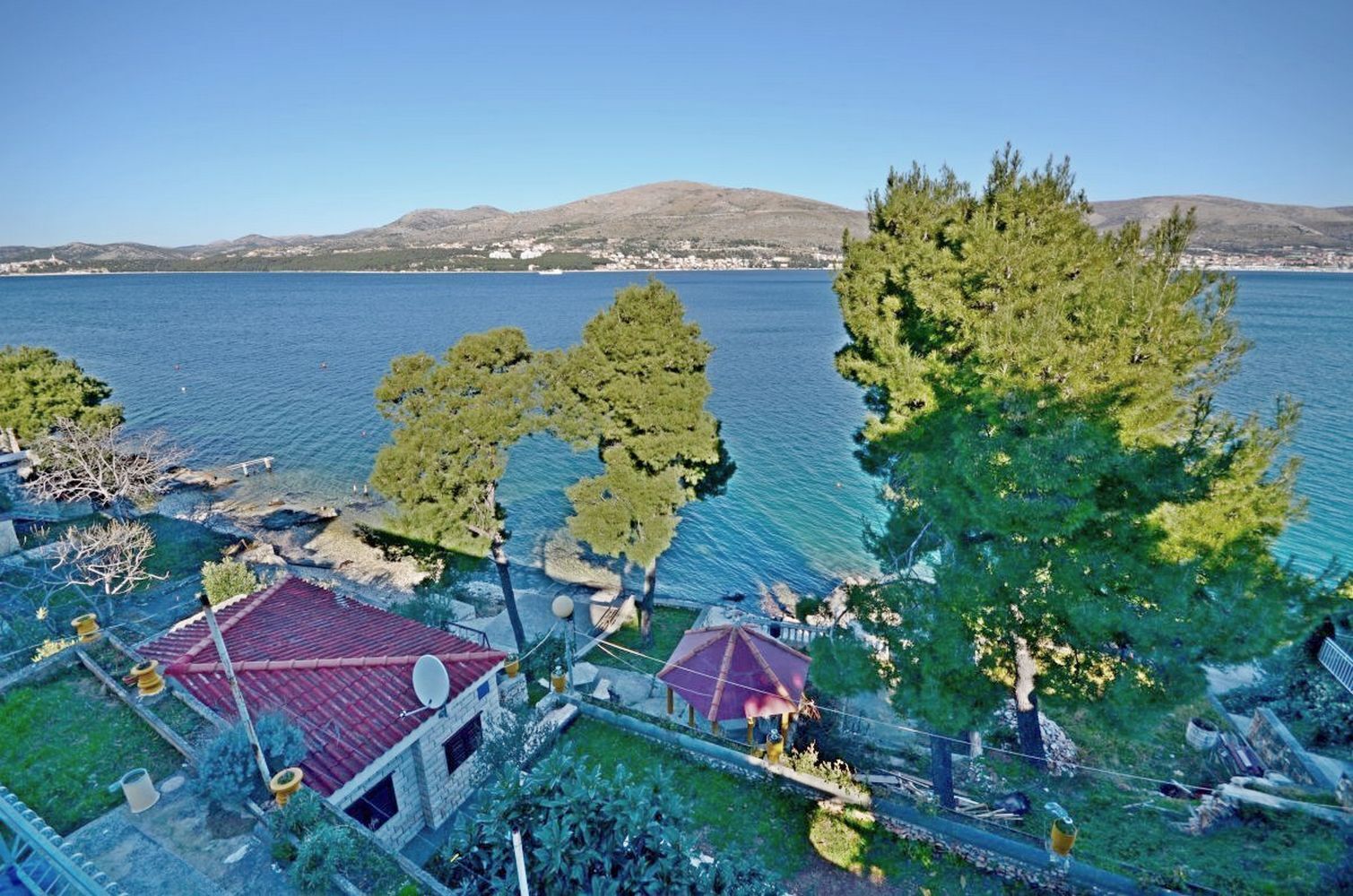 A coastal property with a red-roofed building, a garden, and a view of the sea and distant hills.