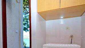 Une petite buanderie équipée d’un lave-linge à chargement frontal, d’un meuble en bois placé au-dessus, et offrant une vue sur un jardin grâce à une porte ouverte.