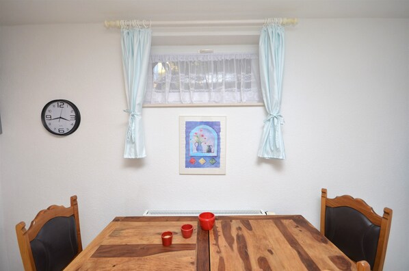 A dining area with a wooden table, two chairs, a clock, and a window with curtains. - Apartments Mara (Vodice)