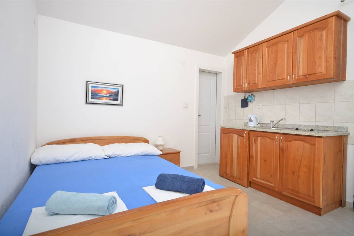 Ein Schlafzimmer mit einem Bett, einer blauen Bettdecke, einem weißen Kopfkissen und einem zusammengerollten Handtuch. Es gibt einen Holzschrank mit einem Waschbecken und einem Wasserkocher. Ein gerahmtes Bild hängt an der Wand.