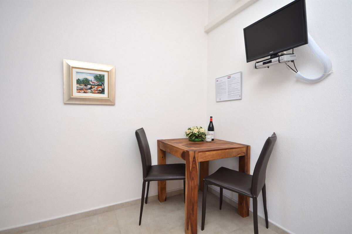 Ein kleines Zimmer mit Holztisch, zwei Stühlen, einem gerahmten Bild, einem angebrachten Fernseher und einer kleinen Pflanze mit einer Flasche auf dem Tisch.