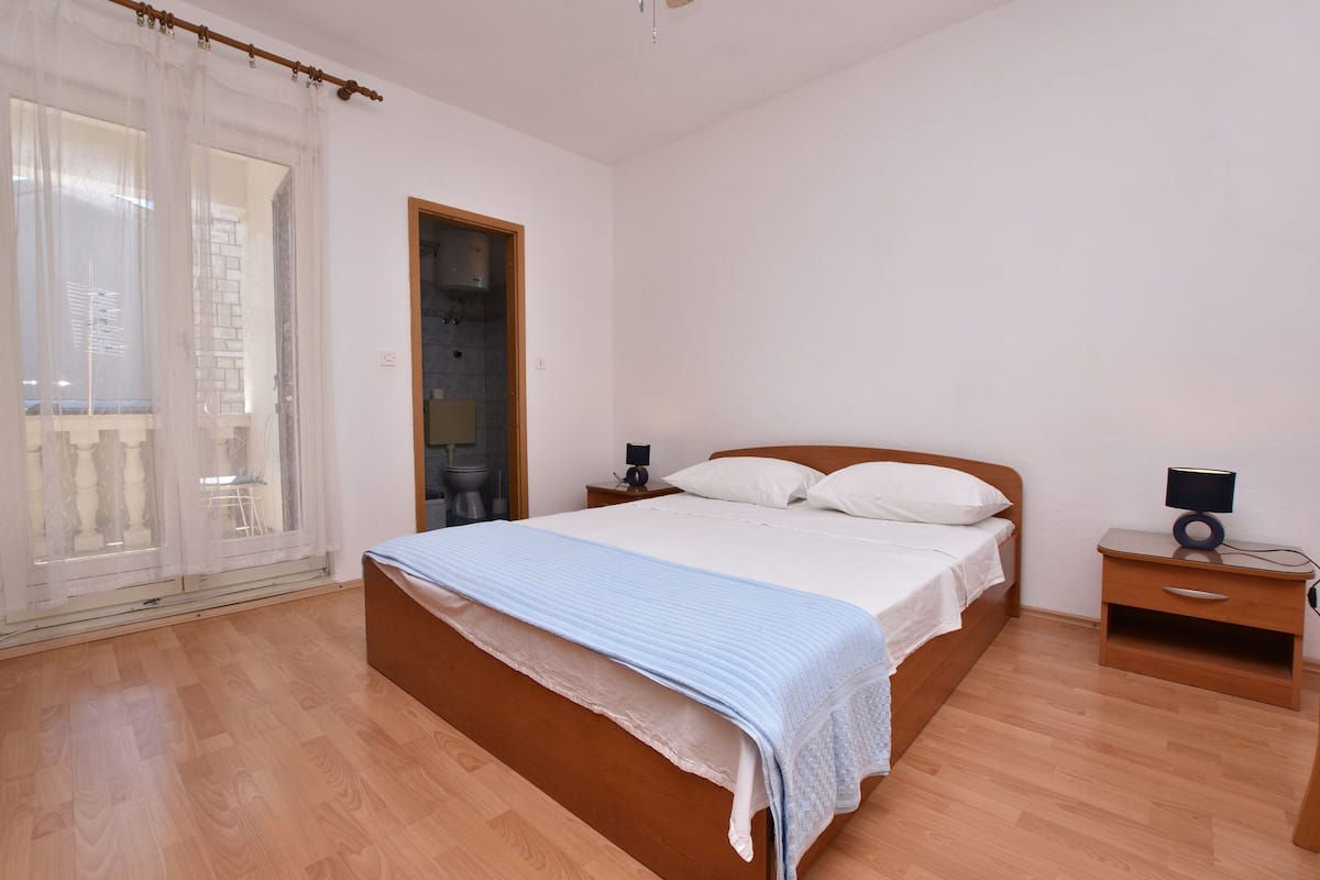 Un dormitorio con cama de madera, una mesita de noche, una lámpara y un ventanal con cortinas de gasa.