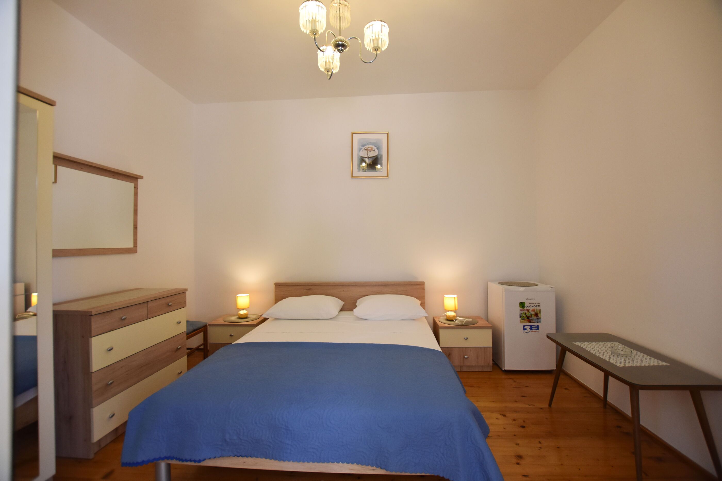A bedroom with a bed, bedside tables, a dresser, a ceiling light fixture, and a refrigerator.