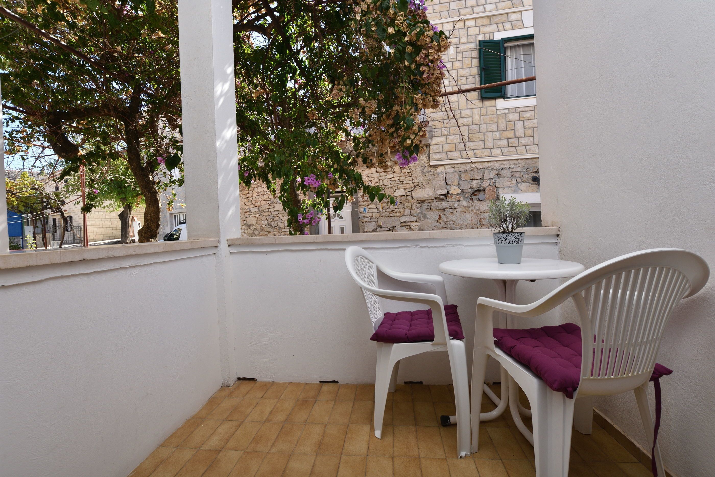 A balcony with a white table and two white chairs, each with a purple cushion. A tree with purple flowers is visible through the window.