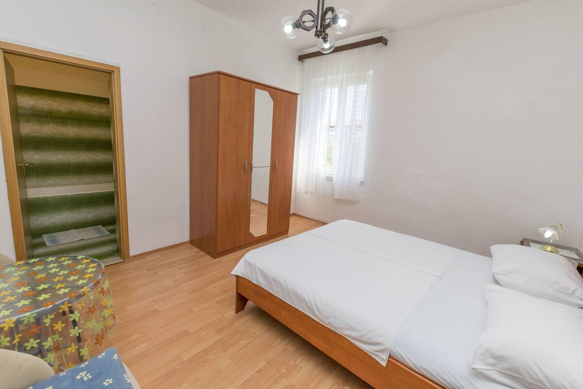Un dormitorio con una cama de madera, un armario, una mesita redonda y una ventana con cortinas de encaje.