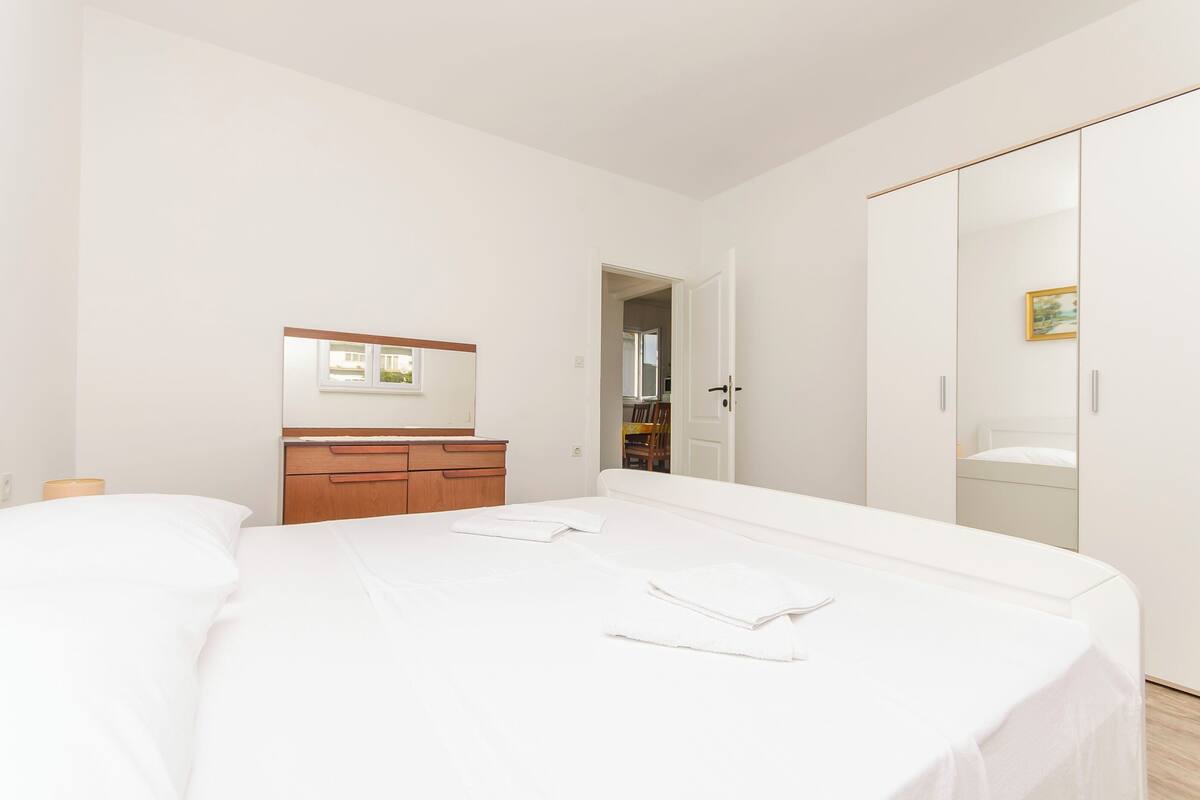 A bedroom with a large bed, a wooden dresser, a wardrobe, and a mirror on the wall.