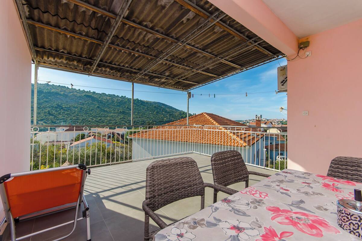 A balcony with wicker chairs and a table, overlooking a mountainous landscape and a red-roofed building.