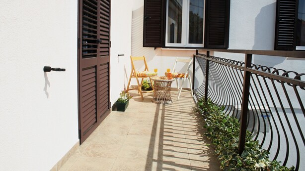A small outdoor patio with a table and chairs, a window with shutters, and a metal railing. - Apartment Anton (Slatine)