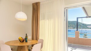 A dining area with a round wooden table, white chairs, a hanging light fixture, and a view of a coastal landscape through large windows. - Apartment Danijela (Primosten)