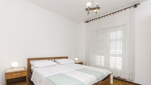 A bedroom with a wooden bed, a bedside table with a lamp, and sheer curtains on the window. - Apartment Niko (Vodice)
