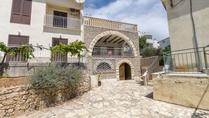 A stone-paved courtyard with a central archway, stone wall, and a building with balconies and shutters. - Apartment Smiljan (Primosten)