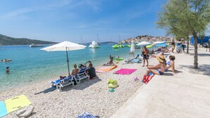 A pebbled beach with lounge chairs and umbrellas, a clear blue sea, and boats in the background. - Apartment Smiljan (Primosten)