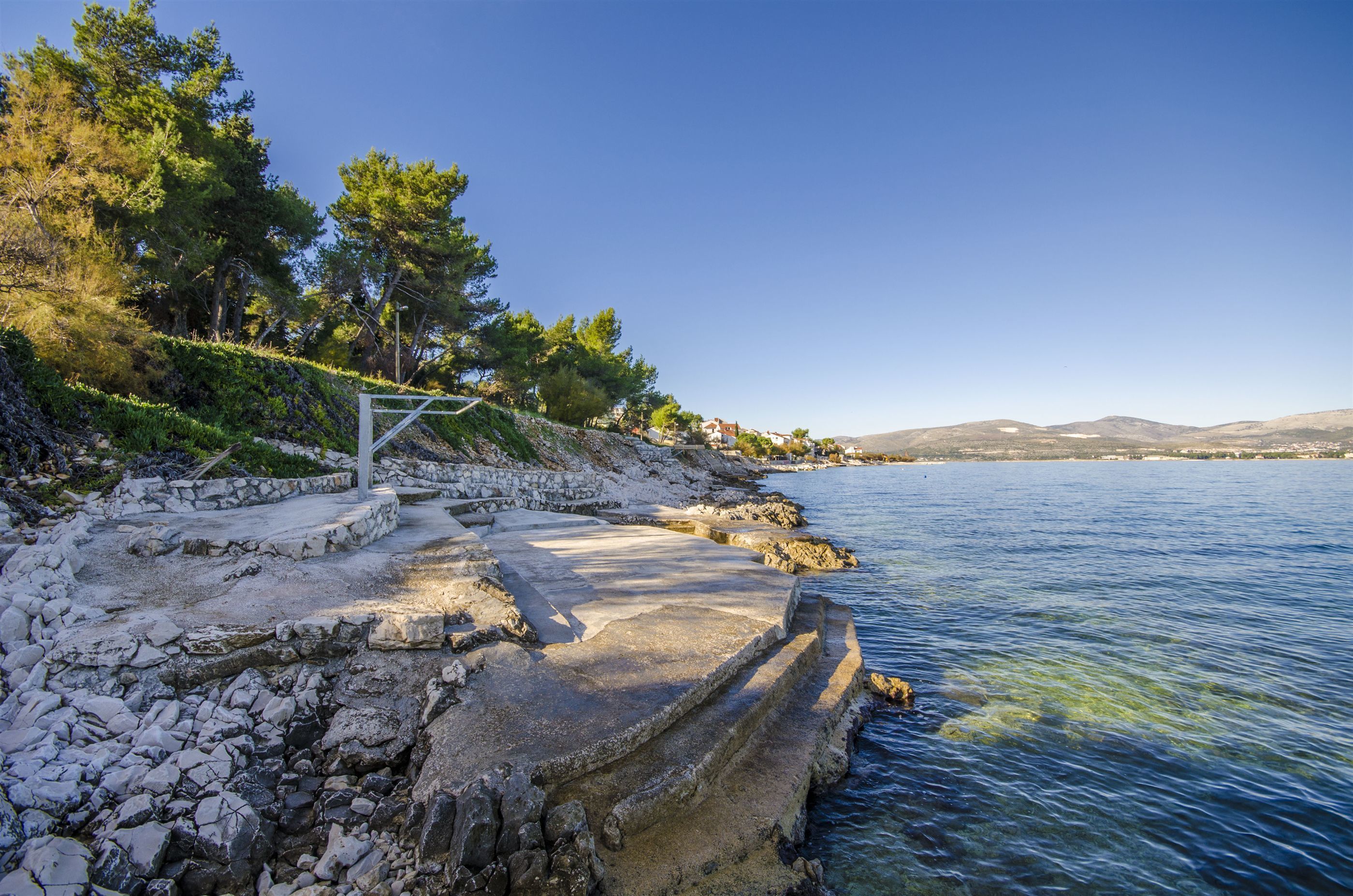 A coastal area with a stone pathway leading to a body of water, surrounded by trees and hills in the background.