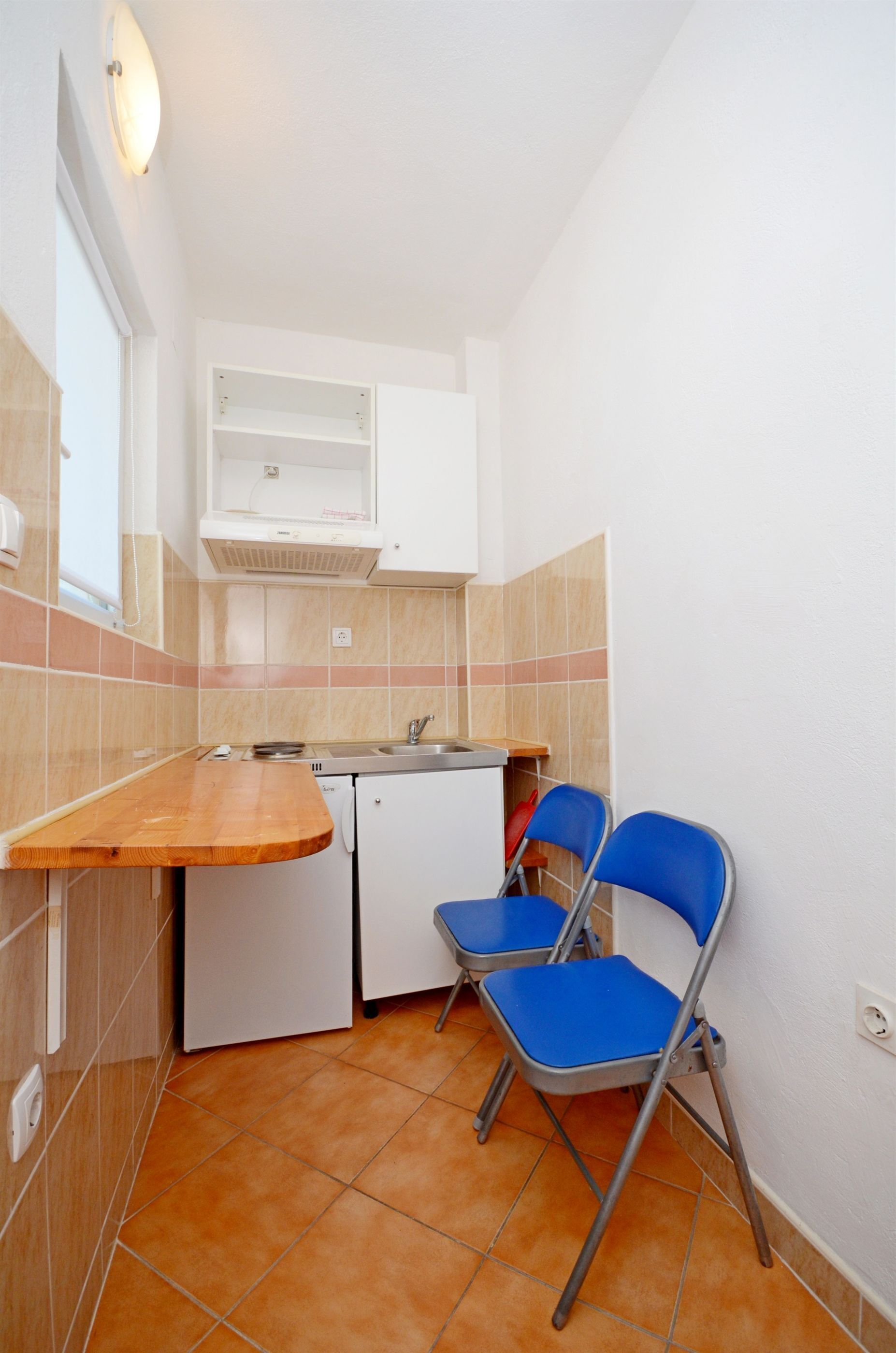 A compact kitchen with a wooden countertop, a sink, a white refrigerator, and a blue folding chair.