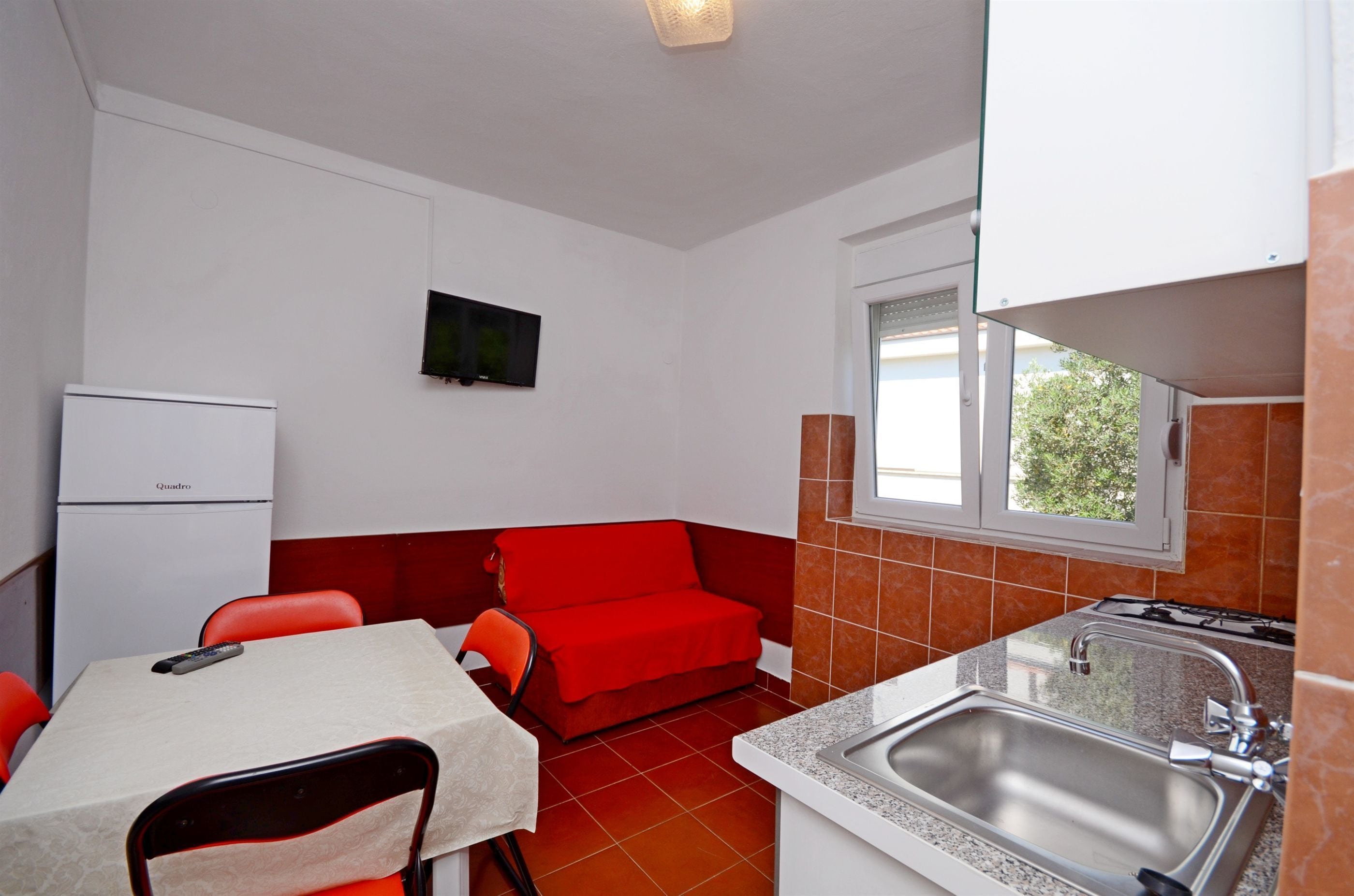 Cocina compacta con refrigerador, una mesa de comedor con sillas, un sofá rojo, una ventana con vistas, y un lavamanos.