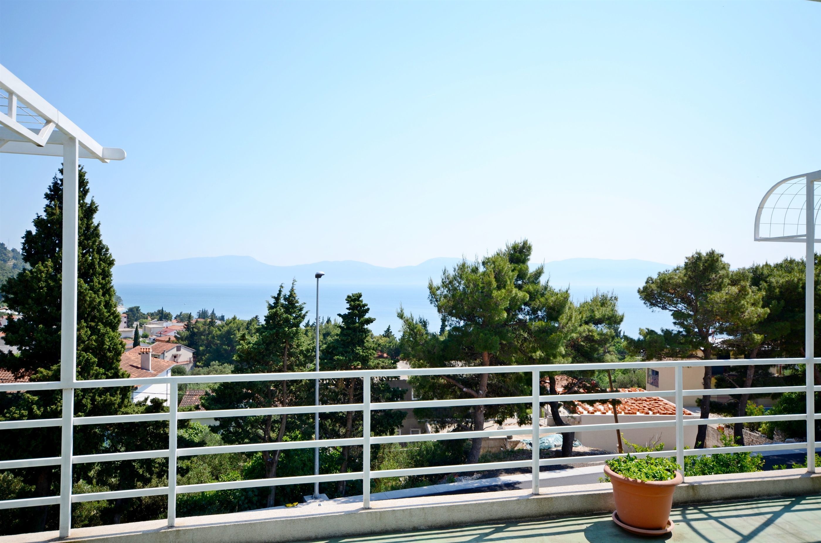 Ein Balkon mit weißer Brüstung, einem Blumentopf und Blick auf eine Stadt und Berge.