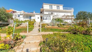 A two-story building with a balcony on the upper floor, a stone pathway leading to the entrance, and a garden with various plants and a trellis. - Apartments and Room Milka-jere (Primosten)