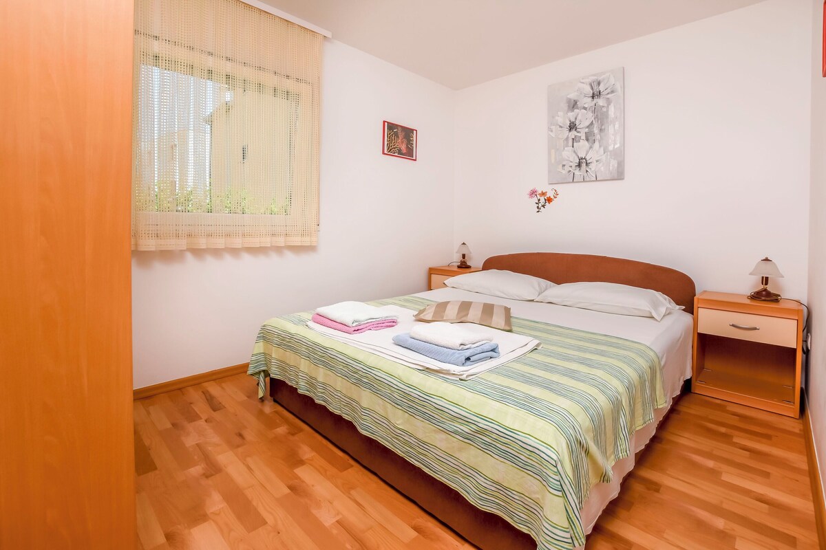 Un dormitorio con cama, piso de madera, una ventana con persianas, una mesita de noche y un cuadro en la pared.