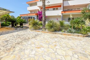 A modern building with a stone-paved courtyard, surrounded by various plants and a palm tree. - Apartments Aurel (Rogoznica)