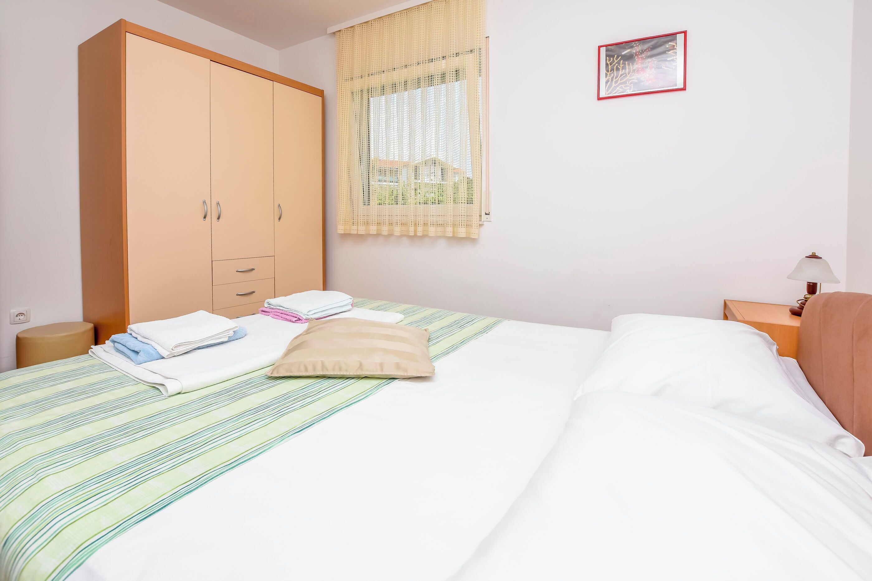 Uma cama bem arrumada com colcha listrada, um guarda-roupa espaçoso, uma mesa de cabeceira com abajur e uma janela com vista para uma casa.