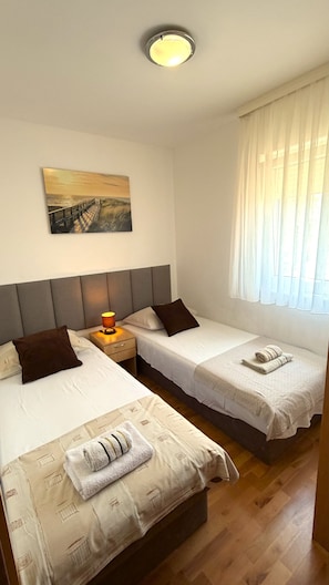 A hotel room with two beds, a nightstand with a lamp, a painting on the wall, and a window with curtains. - Apartments Aurel (Rogoznica)