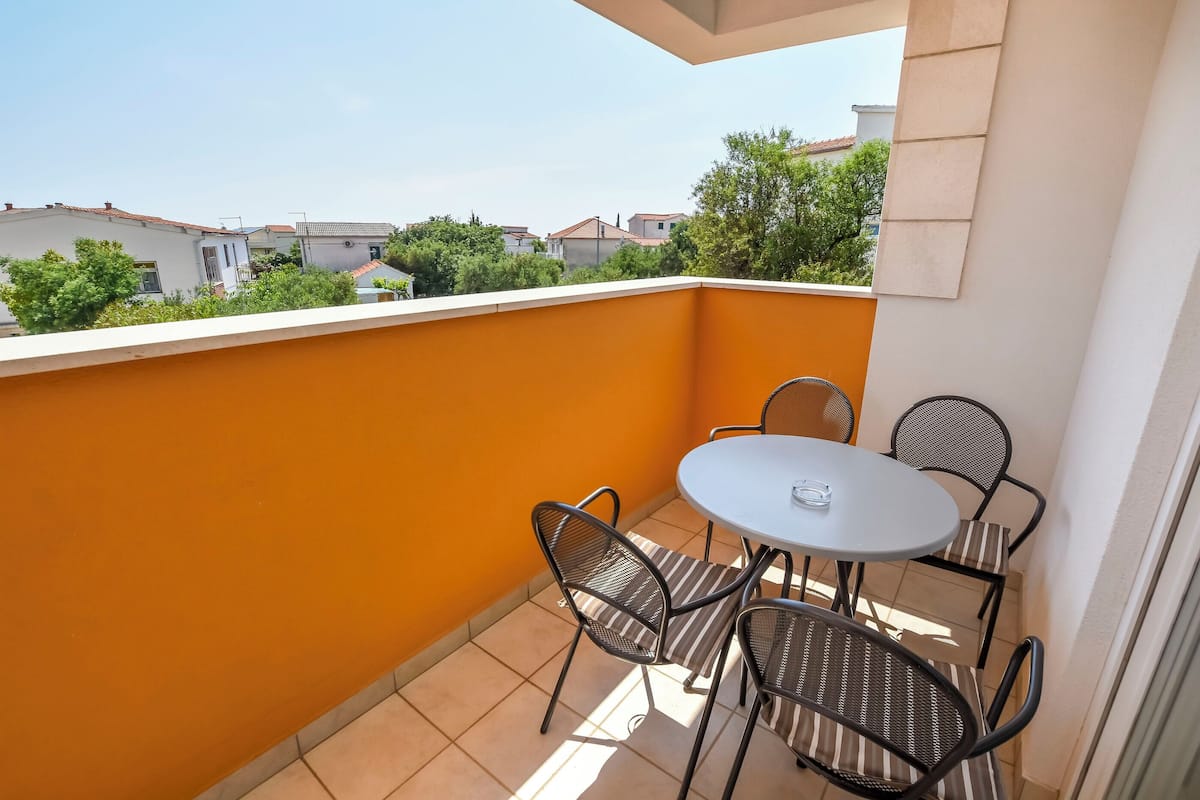 A balcony with a round table and four chairs, a tiled floor, and a view of neighboring houses and trees.