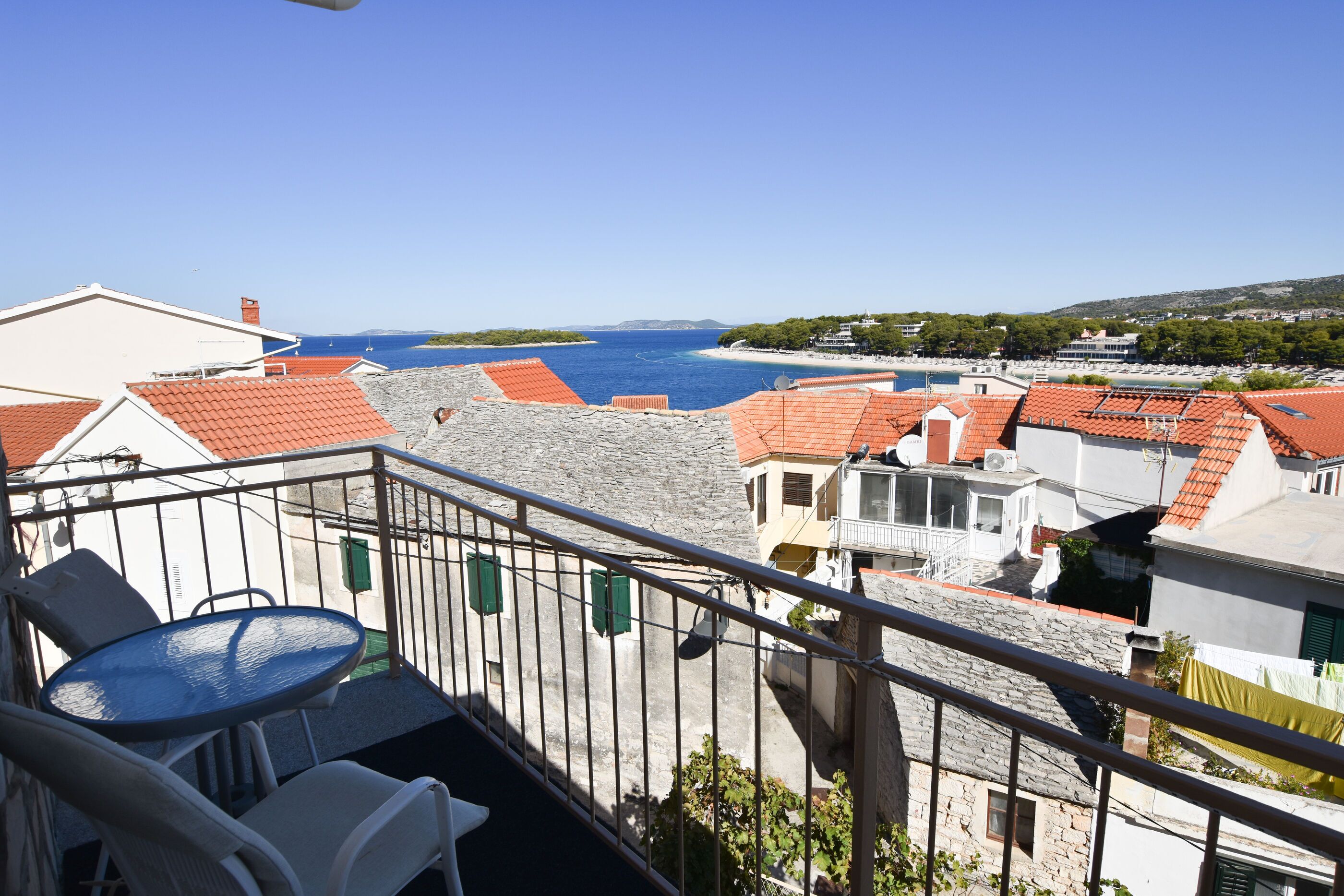 Un balcón con mesa y sillas que da a una villa costera con edificios de techo rojo y un mar azul cristalino.