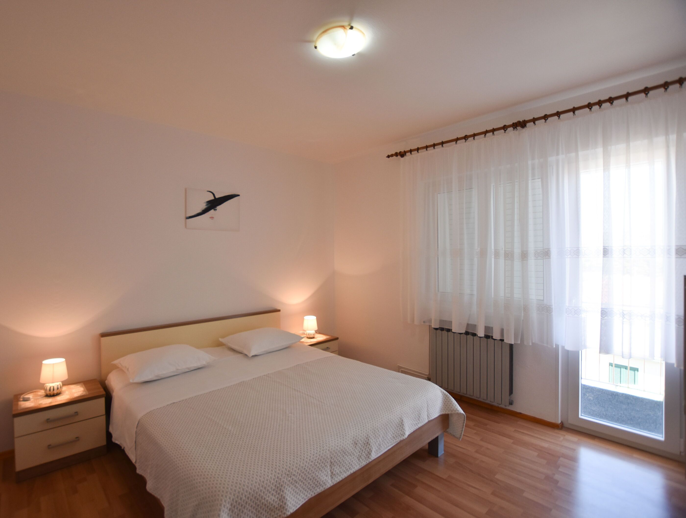 A bedroom with a bed, bedside tables, a lamp, a wall art piece, and a window with curtains.