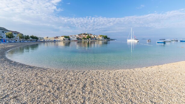 A pebbled beach with clear water, boats, and buildings in the background. - Apartments Zdenka (Primosten)