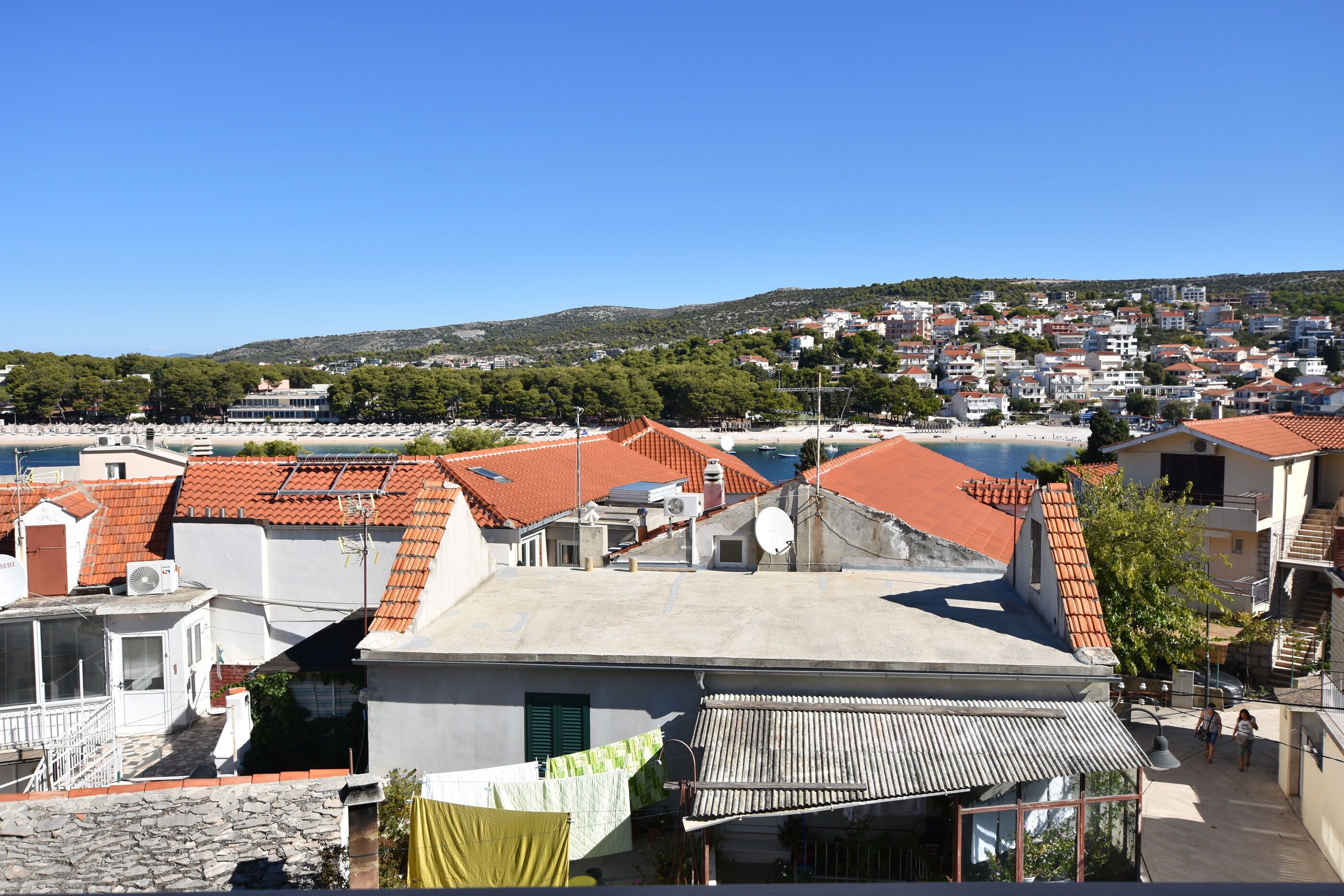 Une ville côtière aux bâtiments aux toits de tuiles rouges, sous un ciel bleu azur, avec des collines en arrière-plan.