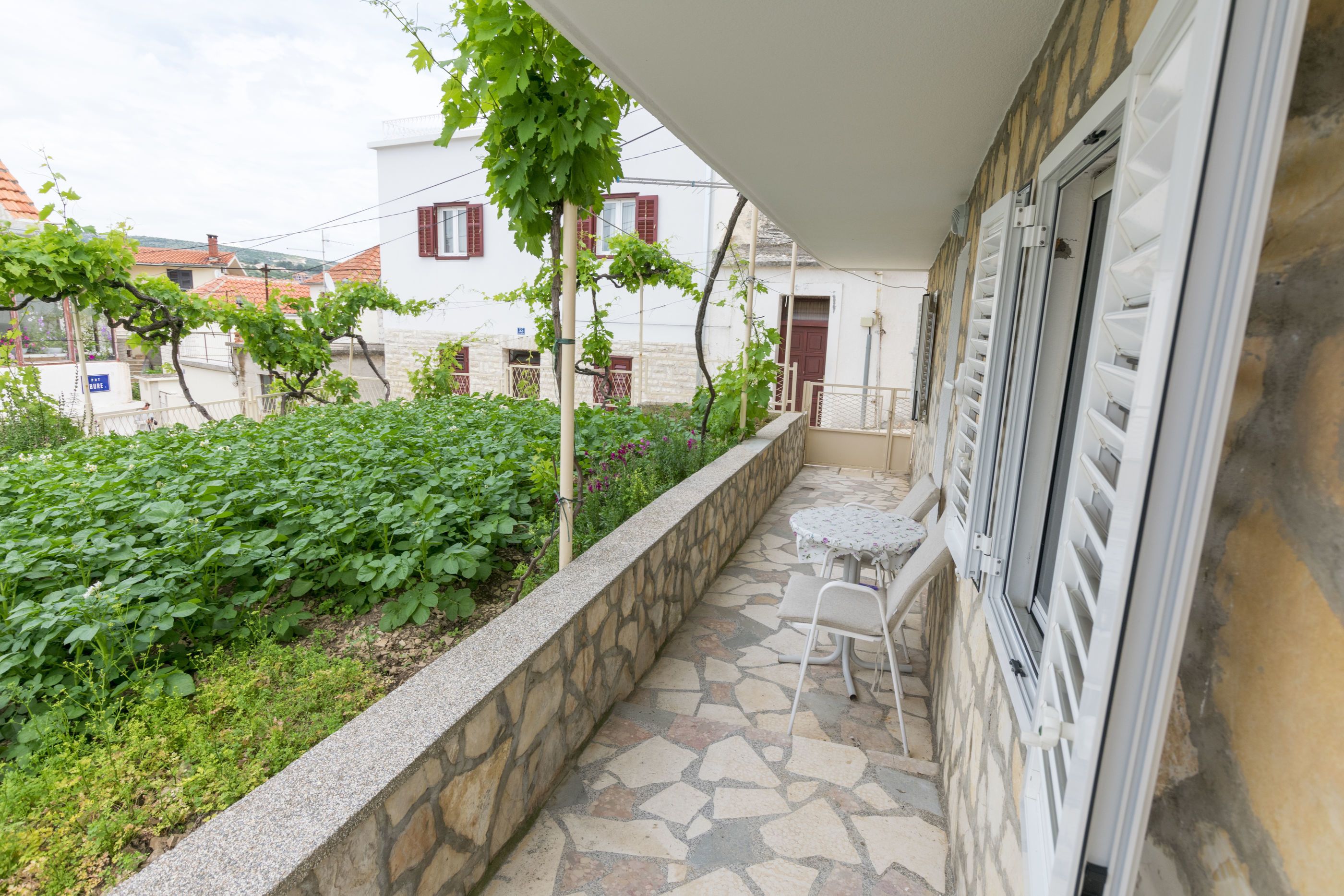 A covered outdoor area with a stone floor, a small table and chairs, and a view of a garden with plants and a building in the background.